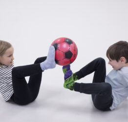 Two children facing each other with their legs in the air. They are holding a red football up with their feet.