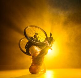 A dancer in a wheelchair performing a head stand in front of a yellow light.