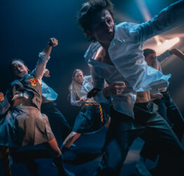 Five dancers in school uniforms, throwing their left arms up in the air with clenched fists.