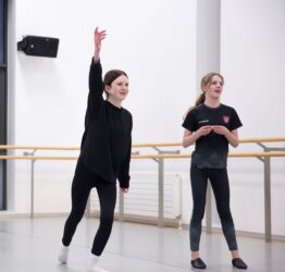 Two young dancers in a studio. One has their right hand in the air and their right foot pointed behind them.