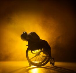 A dancer in a wheelchair leaning backwards against a yellow background.