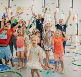 A group of children and three adults in a studio space. Feathers are in mid air and everyone is jumping with their hands in the air.