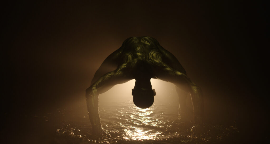 A person bent over looking between their legs. A dark background with water on the floor.