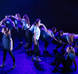 A group of high school students on a stage. A group of them are connected together to create a wave.