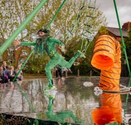 A circular stage outside. A dancers dressed as green algae is midrun. There is an orange pipe in the centre of the stage.