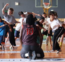 A group of dancers in a sports hall. There is a person knelt on the floor facing them.